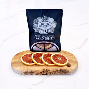 May include: A black bag of "Dried Harvest Co. Premium Cocktail Garnishes" sits atop a wooden oval board with four dried orange slices. The bag features a logo with citrus fruits and leaves.