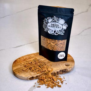 May include: A black bag of Dried Harvest Co. cinnamon sticks with a clear window, displayed on a wooden cutting board. The bag has a white label with the words "Aromatic Stick". Cinnamon sticks are scattered on the board.