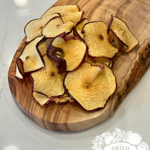 May include: Dried apple slices with reddish-purple edges are arranged on a wooden cutting board. The board has a natural wood grain pattern and is light brown. The image also includes a logo that says "Dried Harvest Co."