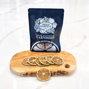 Peut inclure: Un sachet bleu foncé de garnitures à cocktails « Dried Harvest Co. » est posé sur une planche à découper en bois avec des tranches de citron vert séchées. La planche est ovale avec un grain de bois naturel et un petit trou.