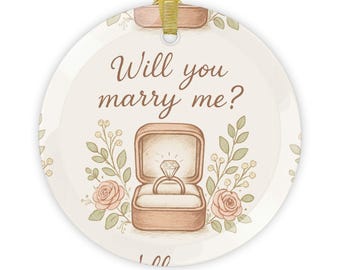 Op de afbeelding: Ronde keramische ornament met een gouden lint om op te hangen. Het ornament toont de tekst "Will you marry me?" boven een tekening van een ring in een doos, omgeven door bloemmotieven in zachte kleuren.