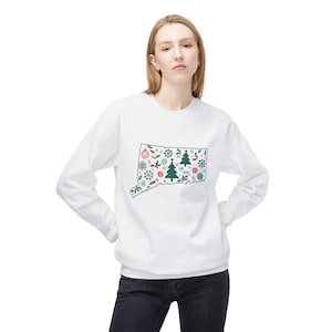 May include: White sweatshirt featuring a green outline of the state of Connecticut filled with Christmas-themed designs. The design includes snowflakes, Christmas trees, and other festive elements. The sweatshirt is a long-sleeved crew neck.
