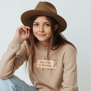 May include: A person wearing a brown fedora hat and a beige sweater with the text "Que Dios te Bendiga" in a floral frame. The person is also wearing blue jeans.
