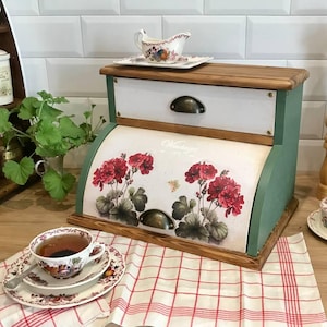 Könnte beinhalten: Eine Brotdose aus Holz mit einer weißen Schublade und einem grünen Rahmen. Die Frontplatte zeigt ein Blumendesign mit roten Blumen und dem Wort "Vintage". Eine Teetasse und Untertasse stehen auf einem rot-weiß karierten Tuch.