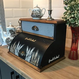 以下が含まれることがあります： 木製の天板とベースが付いた、黒と青のパンボックス。アーチ型の前面には麦の穂と「Fresh Baked」の文字が金色の筆記体で描かれています。パンボックスの上には小さな引き出しがあります。ティーポットと燭台が上にあります。