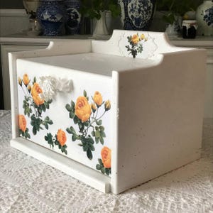 Puede incluir: Panera de madera blanca con un diseño floral de rosas amarillas y hojas verdes. La caja tiene un asa decorativa y un acabado ligeramente envejecido, lo que le da un aspecto vintage. La panera está diseñada para colocarse sobre una encimera.