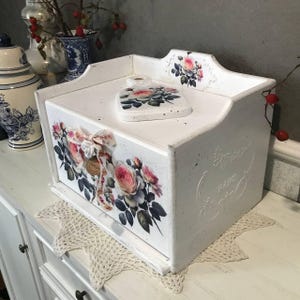 May include: White wooden box with a hinged lid, decorated with floral decals featuring pink roses and green leaves. A matching decorative piece sits on top. The box has a distressed finish and a decorative bow.