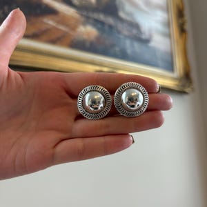 May include: A pair of circular silver-toned earrings featuring a smooth, polished, convex center dome surrounded by a textured, rope-like twisted border. The earrings have a reflective metallic finish and a distinct, layered design.