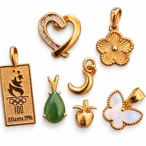 May include: A collection of six gold-toned jewelry pendants including a heart with clear stones, a textured flower, a rectangular Atlanta 1996 Olympic torch charm, a green teardrop stone, a crescent moon, a small apple, and a butterfly with iridescent inlay.
