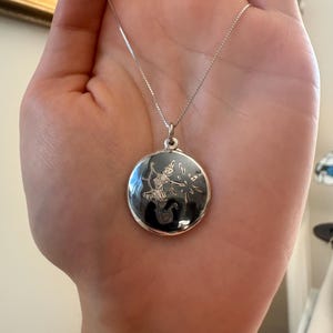 May include: A silver-toned circular pendant necklace featuring a detailed engraving of a figure in a dynamic pose on a dark background. The pendant is suspended from a delicate, thin silver-toned box chain, creating a refined and polished appearance.