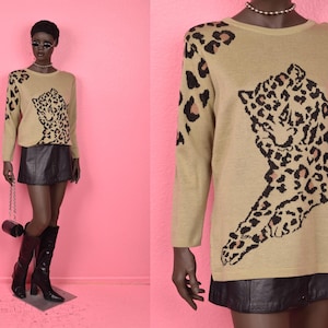 Può includere: Maglione beige a maniche lunghe con un grande disegno di leopardo in nero e marrone. L'outfit include una gonna di pelle nera e stivali neri. L'insieme è presentato su un manichino su sfondo rosa.