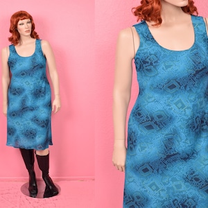 May include: A sleeveless, knee-length dress in shades of blue, featuring a repeating geometric pattern. The dress has a scoop neckline and a slightly flared skirt. The dress is displayed on a mannequin with black boots.
