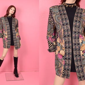 May include: A long, open-front jacket with a floral and paisley pattern in shades of black, gold, pink, and blue. The jacket is worn over a black top and paired with black boots. The mannequin is set against a pink backdrop.