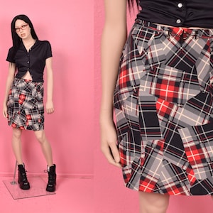 May include: A black, red, and gray plaid skirt. The skirt has a geometric pattern and is paired with a black top. The skirt is knee-length and has a fitted waist. The background is a solid pink.