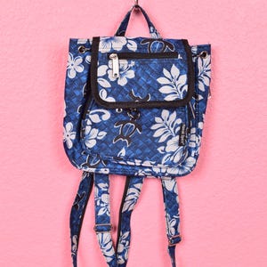May include: Blue backpack with white floral and leaf pattern. Features a front flap pocket, a top zipper, and adjustable straps. The backpack is hanging against a pink background.
