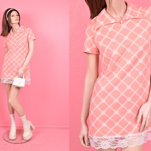 May include: A vintage peach-colored mini dress with a white floral grid pattern and a white lace hem. The dress has short sleeves and a collared neckline. A white handbag is held in the hand.