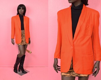 Könnte beinhalten: Leuchtend orangefarbener Blazer mit einem Knopfverschluss und einem fallenden Revers. Der Blazer hat eine Brusttasche mit einem gestickten Design. Die Schaufensterpuppe trägt einen schwarzen Rollkragenpullover, einen Minirock mit Tigerprint und schwarze kniehohe Stiefel.