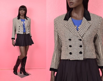May include: A cropped houndstooth blazer with a black velvet collar and trim, paired with a black pleated skirt and knee-high boots. The jacket features black buttons and a double-breasted design. The mannequin is set against a pink backdrop.
