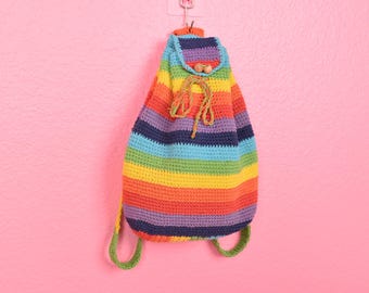 May include: A rainbow striped crocheted backpack with a drawstring closure and wooden beads. The backpack is hanging on a pink wall.