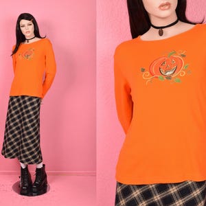 May include: Orange long-sleeve shirt with an embroidered jack-o'-lantern design. The shirt is paired with a black and white plaid skirt. The mannequin is wearing black boots and a black choker necklace. The background is pink.