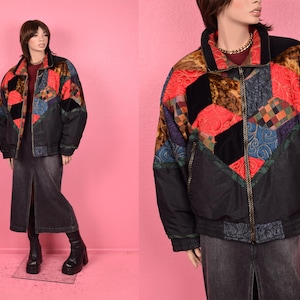 Op de afbeelding: Een zwarte gewatteerde jas met een kleurrijk patchwork-ontwerp met rode, blauwe en bruine geometrische vormen. De jas heeft een ritssluiting aan de voorkant en een kraag. Het model draagt een donkere spijkerbroekrok en zwarte laarzen.