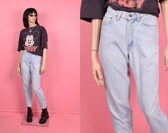 Könnte beinhalten: Hellblaue Jeans mit hoher Taille und Knopfleiste. Die Jeans werden mit einem schwarzen T-Shirt mit Mickey Mouse-Grafik getragen. Das Model trägt schwarze Stiefel und eine Sonnenbrille. Der Hintergrund ist einfarbig rosa.