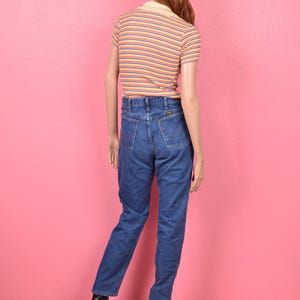 Puede incluir: Un maniqu&iacute; viste un polo a rayas de manga corta con cuello amarillo y vaqueros azules. La camisa tiene rayas naranjas y azules. Los vaqueros son de mezclilla oscura. El maniqu&iacute; lleva botas negras.