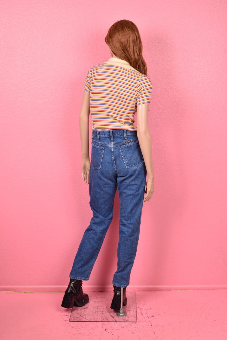 Puede incluir: Un maniqu&iacute; viste un polo a rayas de manga corta con cuello amarillo y vaqueros azules. La camisa tiene rayas naranjas y azules. Los vaqueros son de mezclilla oscura. El maniqu&iacute; lleva botas negras.