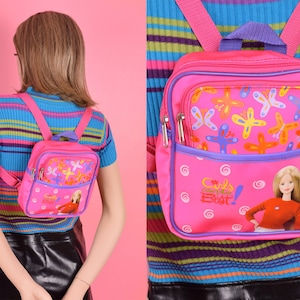 Puede incluir: Mochila rosa con un patrón de mariposas y remolinos. La mochila tiene un bolsillo frontal con una imagen de una muñeca Barbie y el texto "Girls Best!"