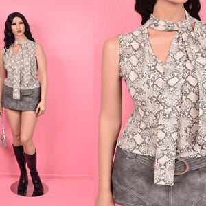 May include: Sleeveless top with a snakeskin pattern in beige and gray, featuring a V-neck and a tie detail. Paired with a gray mini skirt and black knee-high boots. A silver handbag completes the outfit. The mannequin is standing on a reflective surface against a pink backdrop.