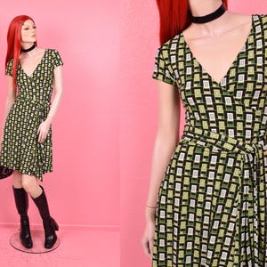 May include: A short-sleeved wrap dress with a geometric pattern in black, green, and white. The dress has a V-neck and a tie at the waist. The mannequin is wearing black boots and a black choker.