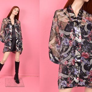 May include: Sheer black floral print kimono-style top with a V-neck and long sleeves. The top features a colorful floral pattern with red, purple, and white flowers. The garment is displayed on a mannequin with a black dress and black boots.