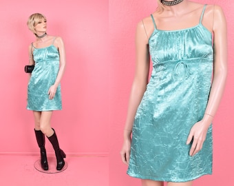 May include: A light teal mini dress with spaghetti straps and a tie detail at the waist. The dress has a textured, crinkled fabric and is displayed on a mannequin. The mannequin is wearing black boots and a black choker necklace. The background is pink.