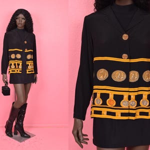 May include: Black blazer with gold coin and geometric designs. The blazer has long sleeves and gold buttons. The outfit is paired with a black skirt and black knee-high boots. A small black purse is held in the hand.