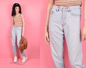 Puede incluir: Vaqueros de mezclilla de color claro con cintura alta y bragueta de botones. Los vaqueros se combinan con una camiseta sin mangas a rayas en tonos beige y melocotón. Una mochila de cuero marrón y zapatillas blancas completan el look.
