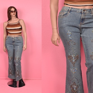 May include: Light wash denim jeans with embellished floral and geometric designs. The jeans have a flared leg and are paired with a brown and tan striped tank top. The mannequin is wearing black boots and sunglasses.