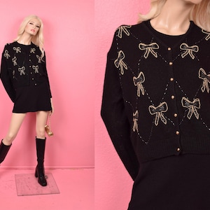 May include: Black cardigan sweater with gold bow embroidery and gold button closures. The sweater is paired with a black dress. The mannequin is wearing black knee-high boots. The background is pink.