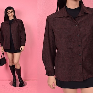 Può includere: Una camicia bordeaux con bottoni e un motivo strutturato. La camicia a maniche lunghe ha un colletto e una tasca sul petto. La camicia è abbinata a una gonna e stivali neri. Il manichino indossa occhiali e tiene in mano una borsa nera.