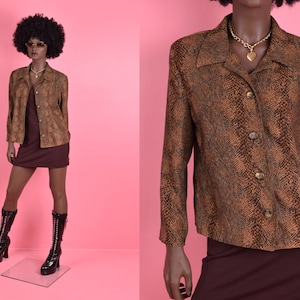 May include: A brown snakeskin patterned button-up shirt with a collar and long sleeves. The shirt has a gold heart-shaped pendant necklace.