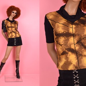 Könnte beinhalten: Ein braunes und gelbes Batik-Shirt mit einem schwarzen Stehkragen und kurzen Ärmeln. Das Shirt wird mit einem schwarzen Minirock mit silbernen Ösen und einem schwarzen Gürtel kombiniert.