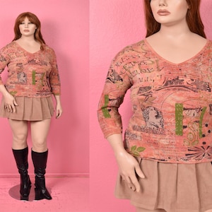 May include: A peach-colored, long-sleeve top with a V-neck and a printed design featuring classical figures and text. The top is paired with a tan pleated skirt and black knee-high boots. The mannequin is set against a pink backdrop.