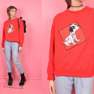 May include: A red sweatshirt featuring a graphic of a Dalmatian dog inside a red diamond. The dog is white with black spots and a black ear. The sweatshirt is paired with light blue jeans and black boots.