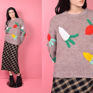 May include: A long-sleeve, crew-neck sweater in a heathered light brown knit featuring embroidered, colorful vegetable motifs in red, white, and yellow with green leafy tops, paired with a black and tan plaid midi skirt and black boots.