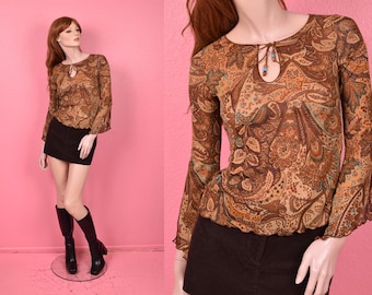 May include: Brown paisley print blouse with long sleeves and a keyhole neckline. The blouse has a flowing, bell-sleeve design. The model is wearing a black mini skirt and black boots. The background is pink.