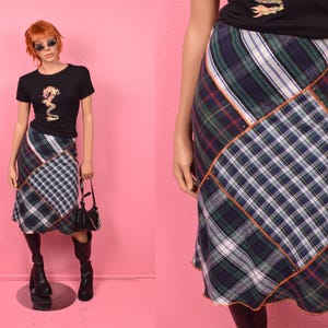 May include: A patchwork plaid skirt in shades of blue, green, and white, with a brown trim. The skirt is paired with a black t-shirt featuring a gold graphic and black boots. The skirt is knee-length.