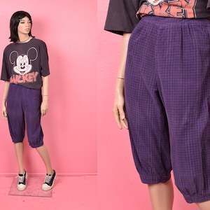 May include: A mannequin wearing a black Mickey Mouse t-shirt and purple plaid capri pants. The capri pants have a button detail on the side and are gathered at the hem. The mannequin is wearing black and white sneakers.