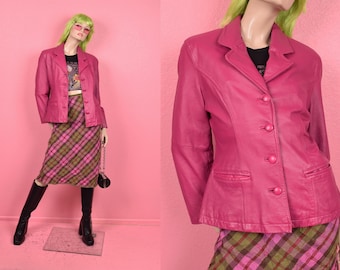 Puede incluir: Una blazer de cuero rosa brillante con tres botones, combinada con una falda a cuadros en tonos rosa, verde y marrón. El conjunto se completa con botas negras y un pequeño bolso negro.