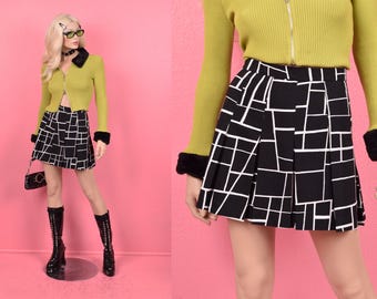 May include: A black and white geometric print mini skirt with a pleated design. The skirt is shown on a model wearing a lime green sweater with black fur trim.
