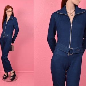 Puede incluir: Un mono de mezclilla azul oscuro con cierre de cremallera, cuello alto y cinturón con una hebilla plateada grande.