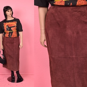 May include: A burgundy suede skirt, knee-length, with a high waist. The skirt is paired with a black graphic t-shirt and black boots. A black handbag is held by the mannequin. The background is a solid pink.